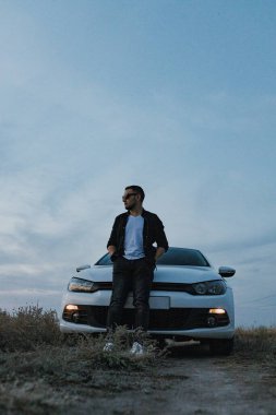 Siyah tişörtlü, kot pantolonlu ve spor ayakkabılı yakışıklı bir adam modern beyaz spor arabanın yanında poz veriyor ve gün batımını seyrediyor. Riverside yakınlarında şehir dışında bir hafta sonu. Koyu renkli fotoğraf