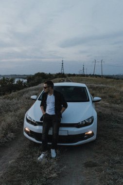 Siyah tişörtlü, kot pantolonlu ve spor ayakkabılı yakışıklı bir adam modern beyaz spor arabanın yanında poz veriyor ve gün batımını seyrediyor. Riverside yakınlarında şehir dışında bir hafta sonu. Koyu renkli fotoğraf