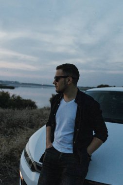 Siyah tişörtlü, kot pantolonlu ve spor ayakkabılı yakışıklı bir adam modern beyaz spor arabanın yanında poz veriyor ve gün batımını seyrediyor. Riverside yakınlarında şehir dışında bir hafta sonu. Koyu renkli fotoğraf