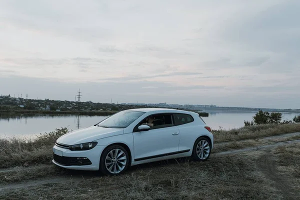 Siyah güneş gözlüklü, beyaz tişörtlü yakışıklı adam modern beyaz spor arabasında oturur ve gün batımını seyreder. Riverside yakınlarında şehir dışında bir hafta sonu. Koyu renkli fotoğraf