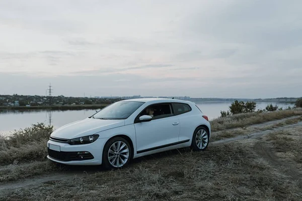 Siyah güneş gözlüklü, beyaz tişörtlü yakışıklı adam modern beyaz spor arabasında oturur ve gün batımını seyreder. Riverside yakınlarında şehir dışında bir hafta sonu. Koyu renkli fotoğraf