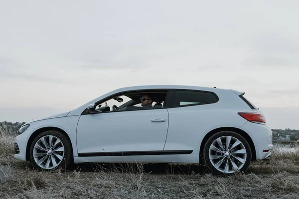 Siyah güneş gözlüklü, beyaz tişörtlü yakışıklı adam modern beyaz spor arabasında oturur ve gün batımını seyreder. Riverside yakınlarında şehir dışında bir hafta sonu. Koyu renkli fotoğraf