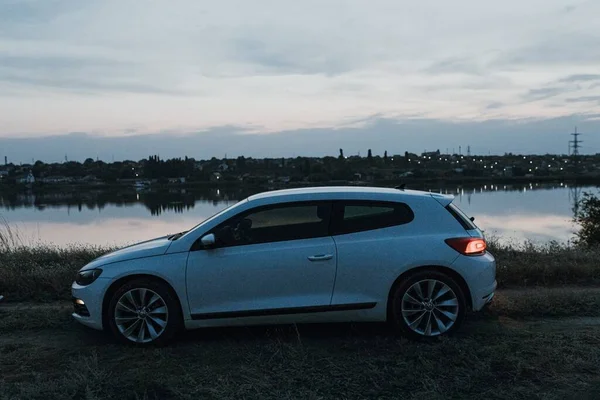 Günbatımında modern lüks beyaz sedan 2 kapılı spor araba. Şehir dışında, kırsal kesimde, nehir kenarında bir hafta sonu. Koyu renkli fotoğraf