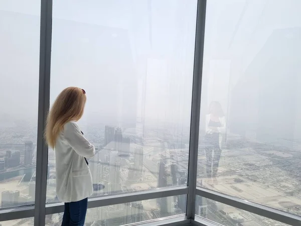 Beautiful girl stands by the window of skyscraper with an amazing panoramic view over the Dubai city and fountains from Burj Khalifa, United Arab Emirates..