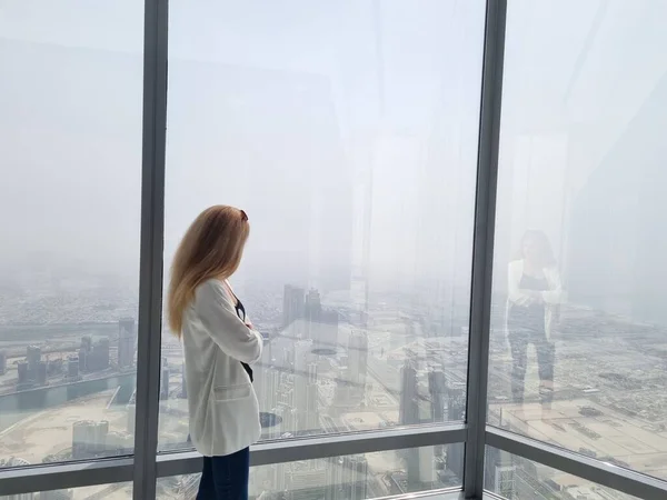 Beautiful girl stands by the window of skyscraper with an amazing panoramic view over the Dubai city and fountains from Burj Khalifa, United Arab Emirates..