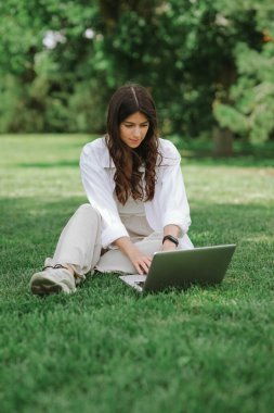 Yaz parkında yeşil çimlerde oturan güzel beyaz kız dizüstü bilgisayarla çalışıyor. Mutlu kadın dışarıda teknoloji, eğitim ve internette sörf yapıyor, sınav konseptine hazırlanıyor.