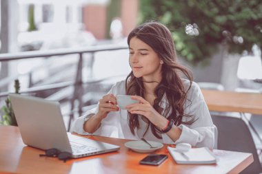 Portre mutlu genç ofis asistanı uzaktan bilgisayarla, tabletle ve kahveyle çalışıyor terasta dizüstü bilgisayarda notlar alıyor ve akıllı telefonu kontrol ediyor. Uzak bağımsız başarılı iş
