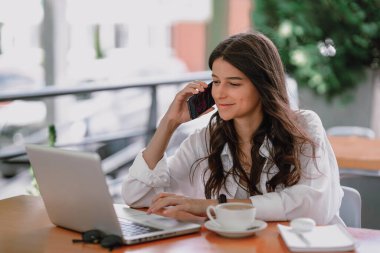 İnternette uzaktan çalışan mutlu genç ofis asistanının portresi. Terasta dizüstü bilgisayarla, tabletle ve kahveyle oturuyor. Aynı anda gündemde notlar alıyor ve telefonda gülümsüyor.  