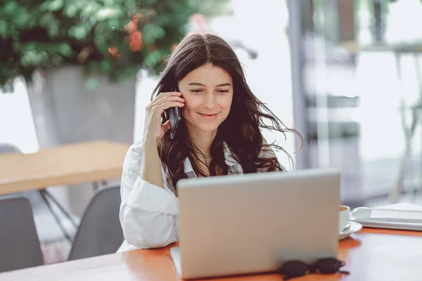 Portrait of happy young office assistant working remotely online sitting with laptop, tablet and coffee outside on terrace making notes in agenda in same time speaking with smile on phone