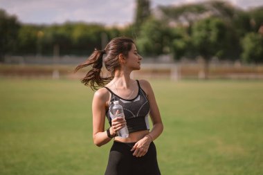 Spor kıyafetli, spor kıyafetli, spor ayakkabılı güzel beyaz kız stadyumun sıcak yazında elinde plastik su şişesi tutuyor. Mutlu kadın açık hava sporu, sağlıklı yaşam tarzı konsepti 
