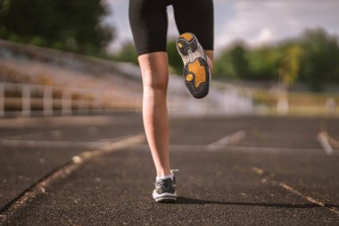 Close-up photo of the Caucasian athlete's leg dressed in sportswear: shorts and top, sneakers with fitness tracker starting running on track at stadium in summer. Happy female outdoor sport concept