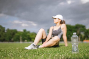 Spor kıyafetleri giymiş, stadyumdaki spor takip cihazıyla beyaz güzel bir kız atlet çimlerde oturuyor. Plastik su şişesine odaklan. Mutlu kadın sporu ve sağlıklı yaşam tarzı konsepti