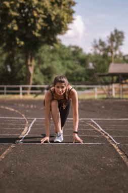 Spor kıyafetleri içinde konsantre beyaz kız atlet şort ve top, spor takip cihazlı spor ayakkabılar yazın stadyumda koşuya hazırlanıyor. Mutlu kadın açık hava sporu konsepti