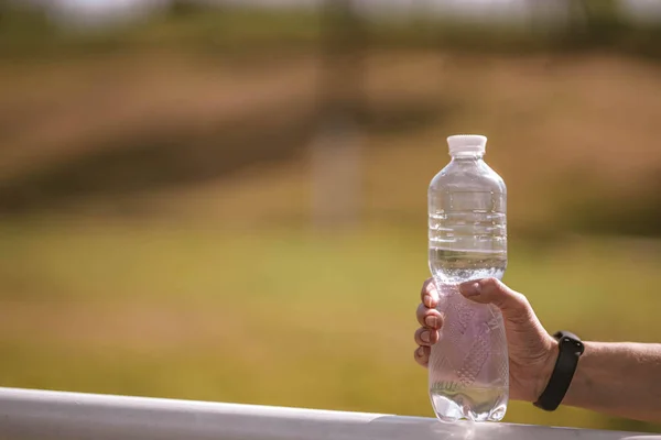 Beautiful Caucasian girl athlete with fitness tracker holds plastic water bottle in her hand on stadium in hot summer. Happy female outdoor sport, healthy lifestyle concept. Blurred  park background