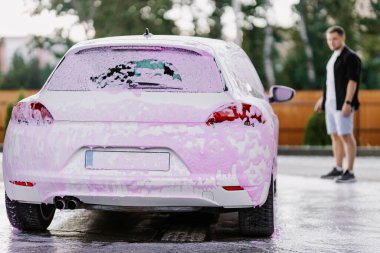 White sedan in pink soapy foam. Contactless self-service car wash. Personal car care cleaning outside. Blurred man. Concept disinfection and antiseptic cleaning of vehicle, covid-19 and coronavirus