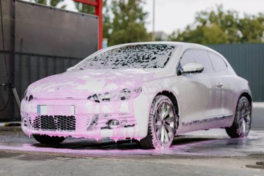 White sedan in pink soapy foam. Contactless self-service car wash concept. Personal car care cleaning outside . Concept disinfection and antiseptic cleaning of the vehicle, covid-19 and coronavirus