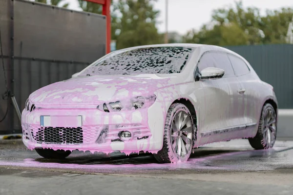 White sedan in pink soapy foam. Contactless self-service car wash concept. Personal car care cleaning outside . Concept disinfection and antiseptic cleaning of the vehicle, covid-19 and coronavirus