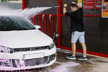 Man washing white car at contactless self-service car wash. Washing sedan car with foam and high-pressure water. Personal car care, cleaning outside. Nozzle spraying a jet of water, process of washing