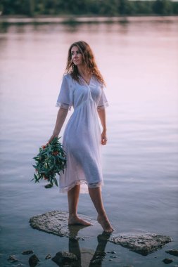 Sensual girl in a white long vintage dress poses on the river stones with a willow and flowers wreath. Harmony and rest with nature. Ukrainian traditional celebration of Ivana Kupala. Quiet place 