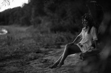 Girl in a white vintage dress with willow and flowers wreath sitting and dreaming near the river on stones . Harmony and rest with nature. Ivan Kupala tradition in Ukraine. Black and white photo