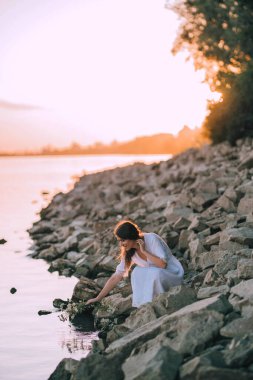 Sensual girl in white vintage dress sits with flower bouquet near the river on stones. Harmony and rest with nature. Ukrainian tradition celebration of Ivana Kupala. Quiet place.