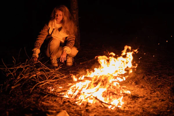 Woman sits near flames fire, remnants bonfire with sparks in forest glade at night near riverside. Charred tree branches. burning out firewood. Big bright flame. Close-up background, selective focu