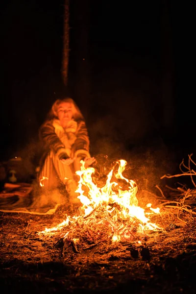Woman sits near flames fire, remnants bonfire with sparks in forest glade at night near riverside. Charred tree branches. burning out firewood. Big bright flame. Close-up background, selective focu