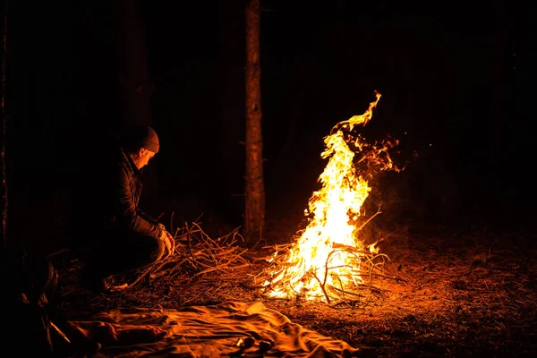 Man sits near flames fire, remnants bonfire with sparks in forest glade at night near riverside. Charred tree branches. burning out firewood. Big bright flame. Close-up background, selective focu