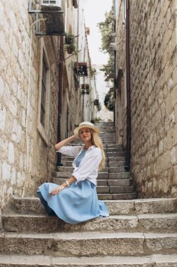 Güzel sarışın bayan turist, mavi elbiseli ve hasır şapkalı gezgin merdivenlerde oturuyor, Hırvatistan 'ın Dubrovnik kentinde dar sokaklar. Eski kasaba 1979 yılında Avrupa 'da UNESCO Dünya Mirasları Listesi olarak listelendi.