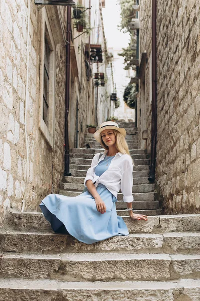 Beautiful blonde lady tourist, traveler in blue dress and straw hat sits on stairs, narrow streets in Dubrovnik, Dalmatia, Croatia. Old town was listed as UNESCO World Heritage Sites in 1979 in Europe