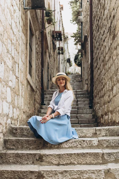 Beautiful blonde lady tourist, traveler in blue dress and straw hat sits on stairs, narrow streets in Dubrovnik, Dalmatia, Croatia. Old town was listed as UNESCO World Heritage Sites in 1979 in Europe