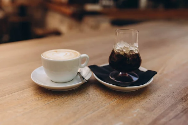 Ice coffee in a tall glass with whiskey and latte coffee in a white cup with a plate on  a wooden table. Cold and hot summer drink on a dark napkin and white plate, light wooden table in cafe