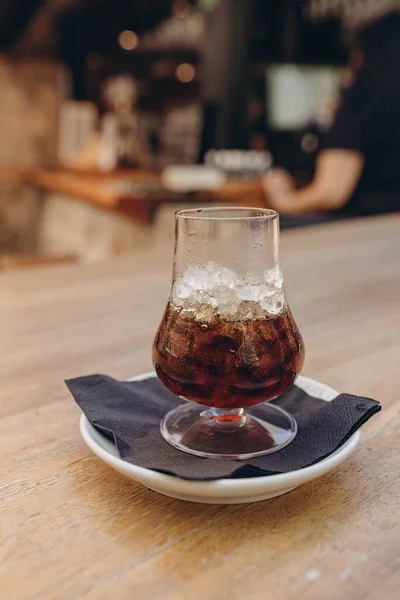 Ice coffee in a tall glass with whiskey and latte coffee in a white cup with a plate on  a wooden table. Cold and hot summer drink on a dark napkin and white plate, light wooden table in cafe
