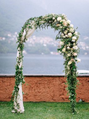 a wedding arch of white roses and greenery, standing on a green lawn overlooking Lake Como, and small towns and mountains