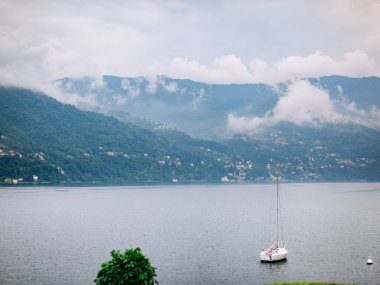 Italy. Lake Como. Lonely white sailing yacht in the water on a background of green hills and small houses and villages in red, yellow, and white colors