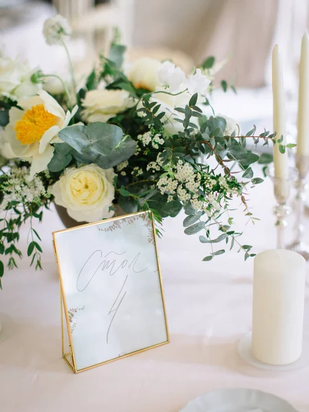 served table, on ivory tablecloths, there is a bouquet of peonies and greenery, candles, and a glass plate with the inscription 