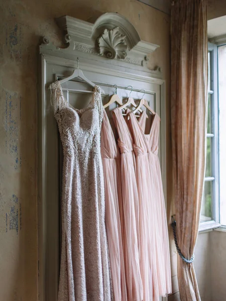 A white wedding dress and bridesmaids\' dresses in pink are hanging on an old vintage wardrobe of light green color.