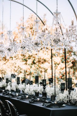 Serving and decoration of the banquet table in black with white flowers, black candles, and crockery. Crystal chandeliers illuminate the table from above.