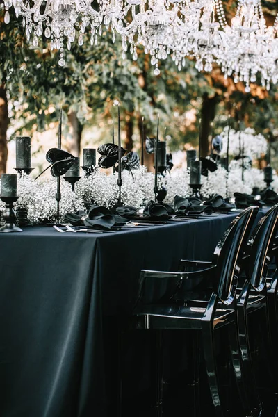 Serving and decoration of the banquet table in black with white flowers, black candles, and crockery. Crystal chandeliers illuminate the table from above.