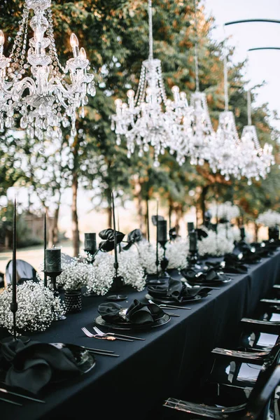 Serving and decorating the banquet table in black with white flowers, black candles, and crockery. Crystal chandeliers illuminate the table from above.