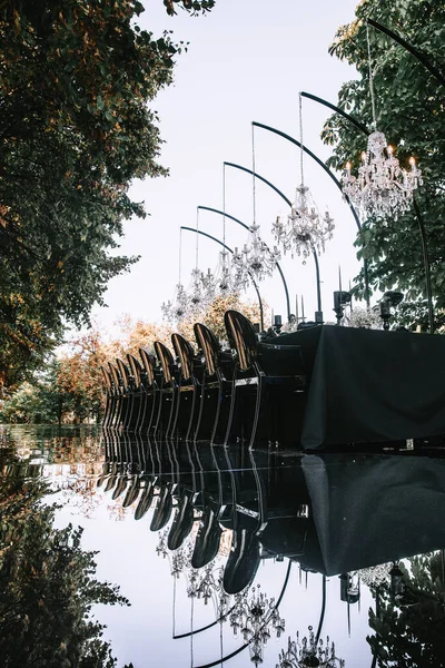 An outdoor banquet table decorated in black with crystal chandeliers on top of a mirrored floor. Reflection on the floor, black and white decor.