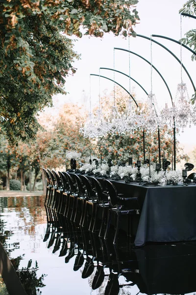 An outdoor banquet table decorated in black with crystal chandeliers on top of a mirrored floor. Reflection on the floor, black and white decor.