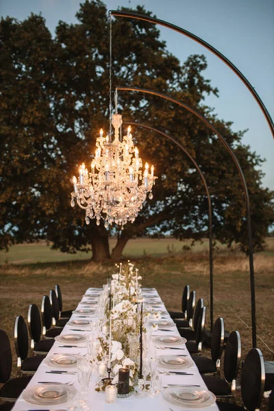 The delicate decor of the wedding banquet table is in pastel colors: wildflowers, crystal glassware, candles. Against the background of a large tree, a crystal chandelier on top, illuminating the table.