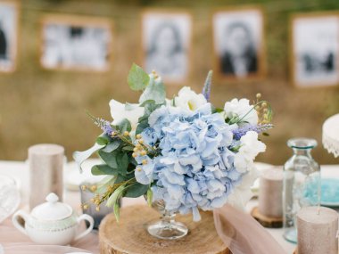 A delicate bouquet of blue hydrangea, white eustoma, lavender, and eucalyptus stands on a wooden stand. In the background are candles and chinaware.