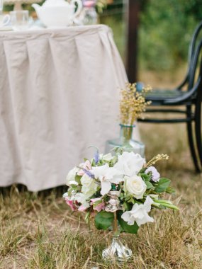 A bouquet of white and lilac eustoma, pink roses, twigs, and leaves stands in a copper candlestick on the grass. In the background is a banquet table with a white tablecloth.