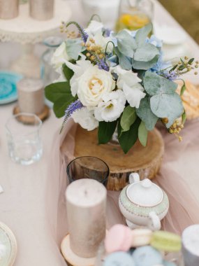 Wedding banquet table decor in pastel colors. A bouquet of eustoma, roses, and eucalyptus stands on a wooden stand. Nearby are candles and dishes.