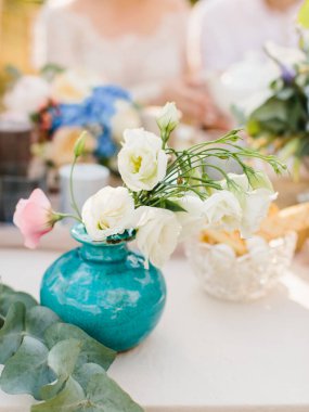 A bouquet of white and pink eustoma stands in a turquoise vase. In the foreground is a branch of eucalyptus.