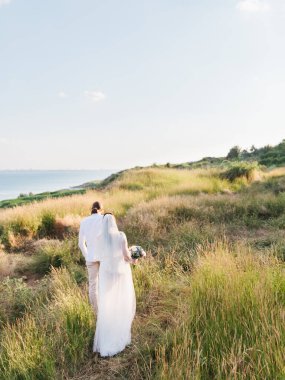 The groom in a beige suit and the bride in a white dress with a long veil is walking along a hill overgrown with steppe grasses. The sea is in the background.