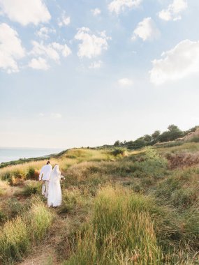 The groom in a beige suit and the bride in a white dress with a long veil is walking along a hill overgrown with steppe grasses. The sea is in the background.
