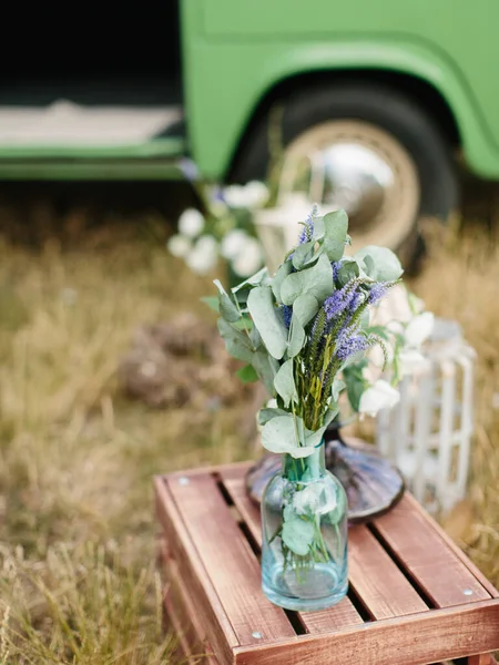A bouquet of eucalyptus and lavender leaves stands in a glass vase on a wooden table outside. In the background is a green Volkswagen hippie van. Outdoor wedding decor.